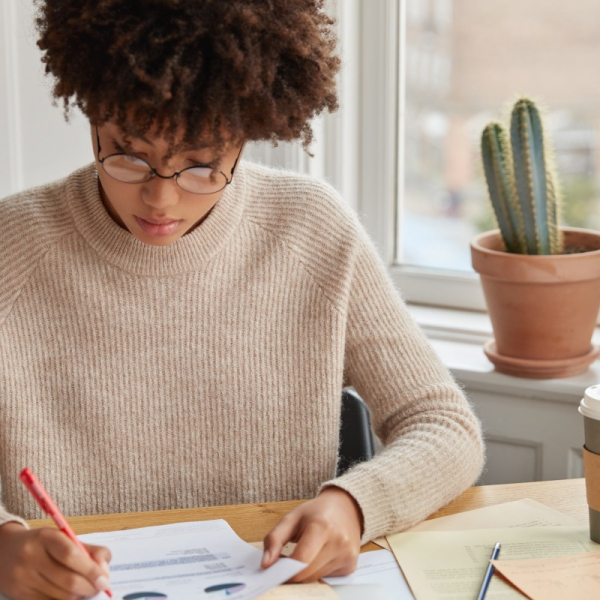 Vertical shot of hard working student with Afro haircut, checks text documentation, prepares pie chart for describing statistics, writes in documents, wears casual sweater. Woman carries accounting
