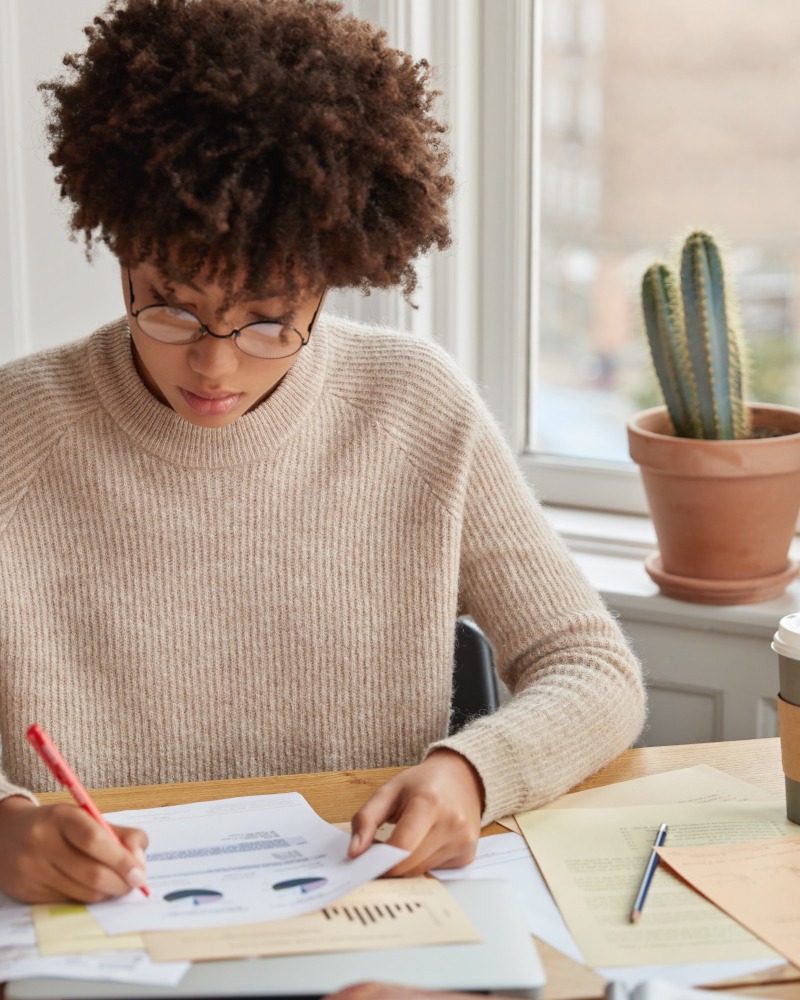 Vertical shot of hard working student with Afro haircut, checks text documentation, prepares pie chart for describing statistics, writes in documents, wears casual sweater. Woman carries accounting